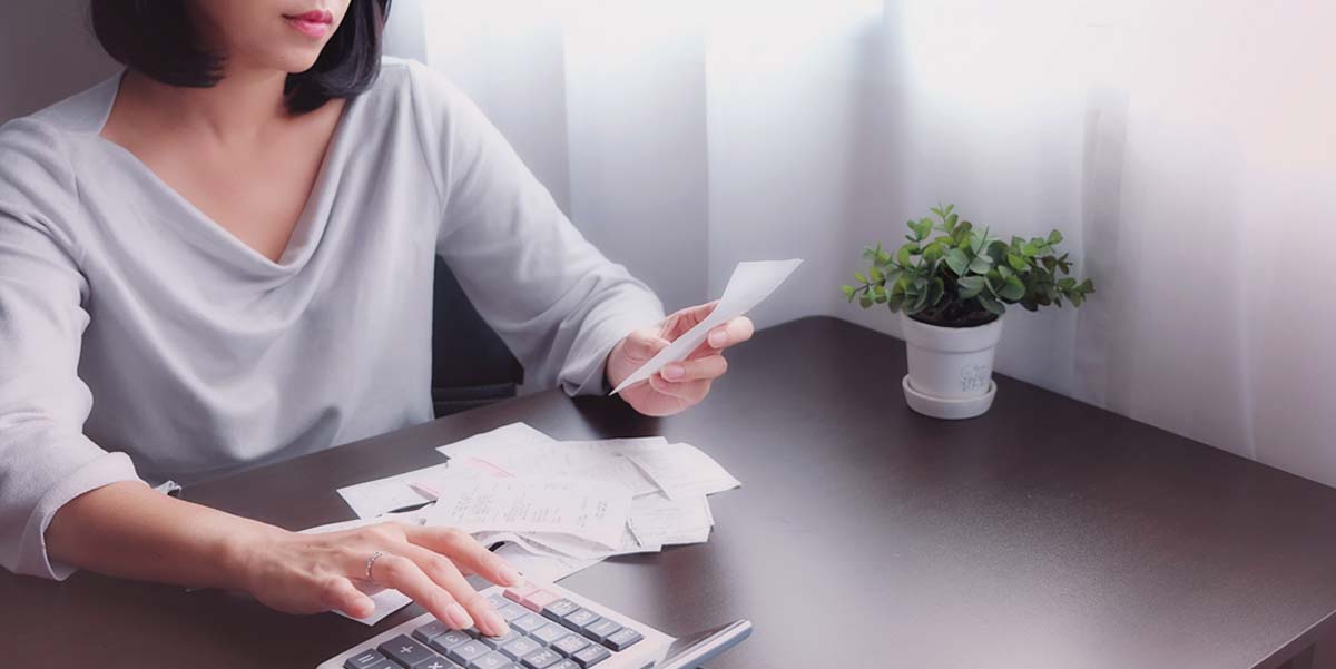 Image: A distressed woman sits at a desk by a window. She holds receipts, papers, and a calculator.