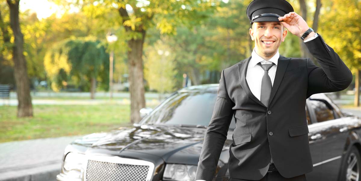 Image: A male Chauffeur smiles in morning light as he stands waiting outside a shiny black limousine.