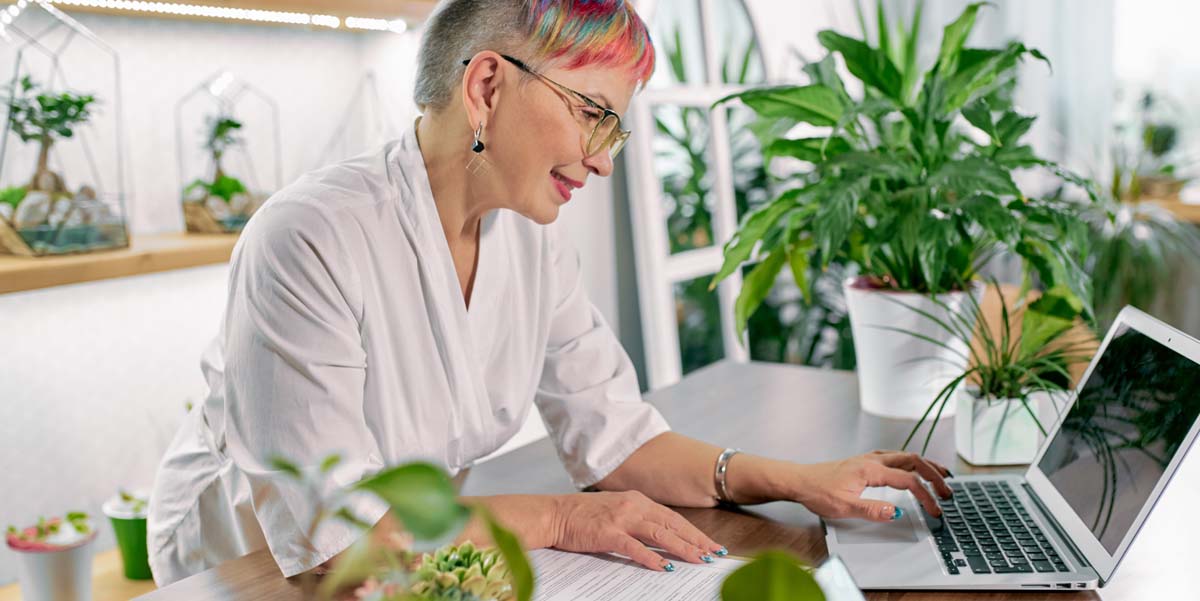 Artistic grey-haired woman sits amongst many plants in plant shop and does paperwork