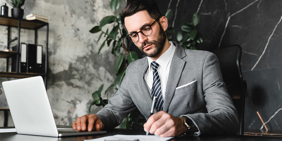 Male account with laptop writing with a pen on a paper document