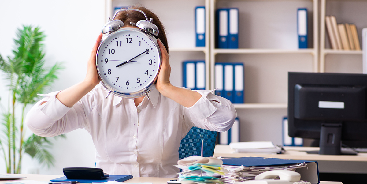 Woman sitting at desk holding a large old-fashioned metal alarm clock in front of her face with both hands