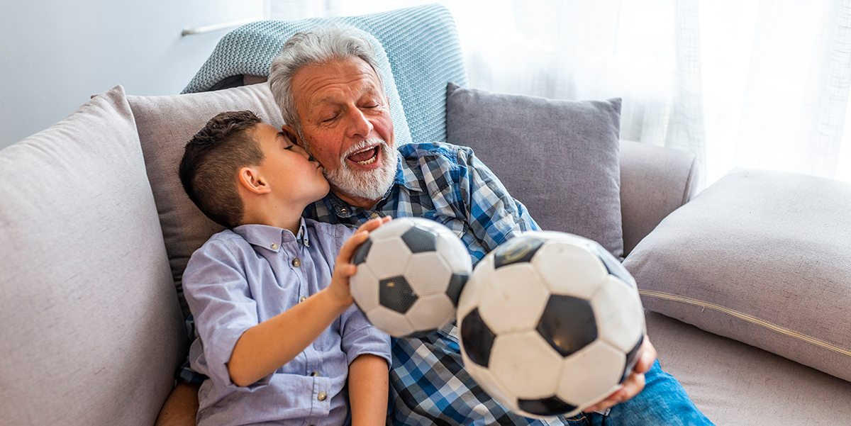 Grandfather with grandson holding soccer ball sitting on the sofa laughing.
