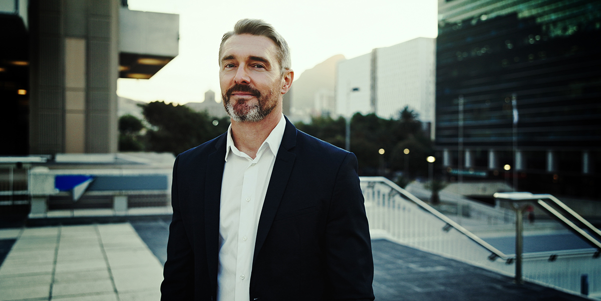 A confident man in an inner-city urban plaza with large buildings in the background.