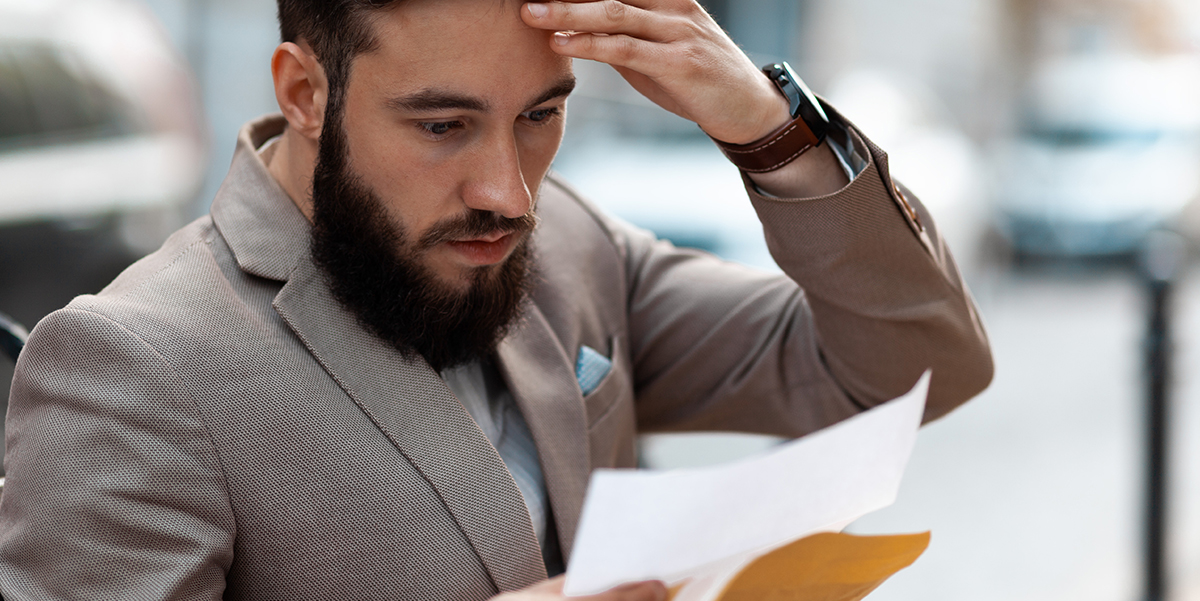 A bearded man with a worried expression is looking at a paper statement and rubbing his forehead.