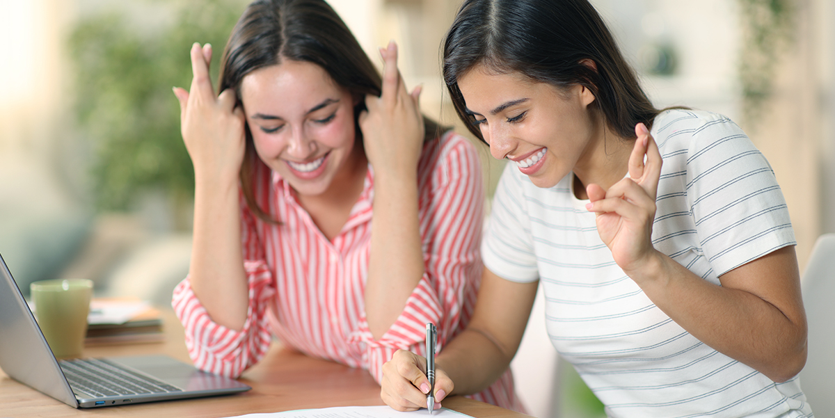 Two young women are looking at a laptop and holding their fingers crossed.
